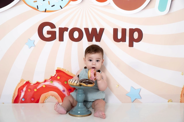 תינוק יושב על רצפה לבנה עם סופגנייה ביד, מול רקע בנושא סופגניות.