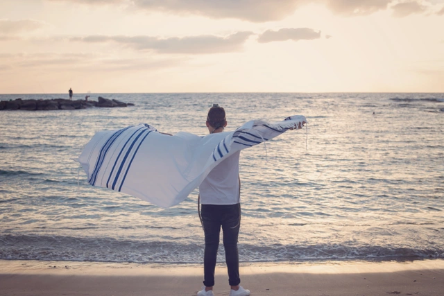 ילד עומד על חוף ים עם טלית פרושה, פונה לים בשקיעה.