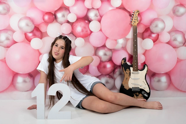 ילדה יושבת על רקע בלוני ורוד עם גיטרה והמספר '12'.