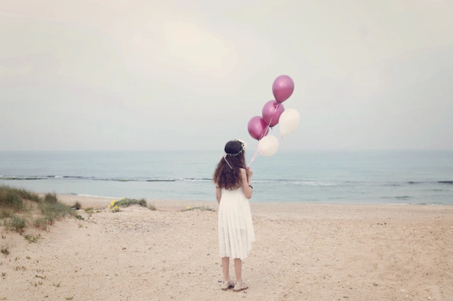 ילדה עומדת על חוף הים, מחזיקה בלונים בצבעי סגול ולבן.