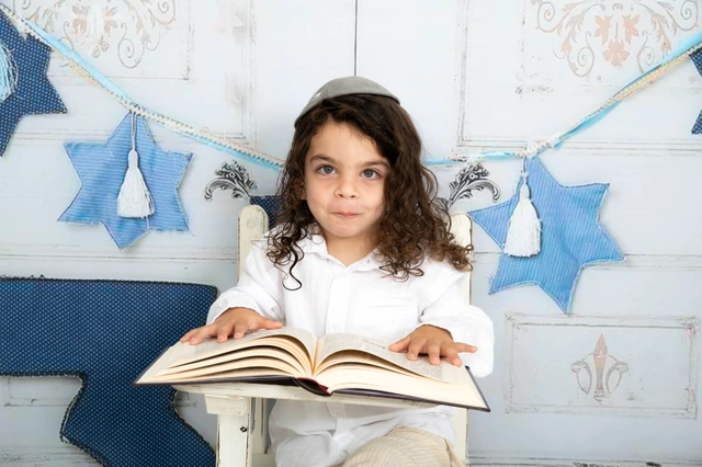 ילד חובש כיפה יושב על כיסא עם ספר פתוח ומביט למצלמה.