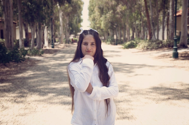 ילדה בחולצה לבנה מצטלמת על שביל עצים, מביטה למצלמה.