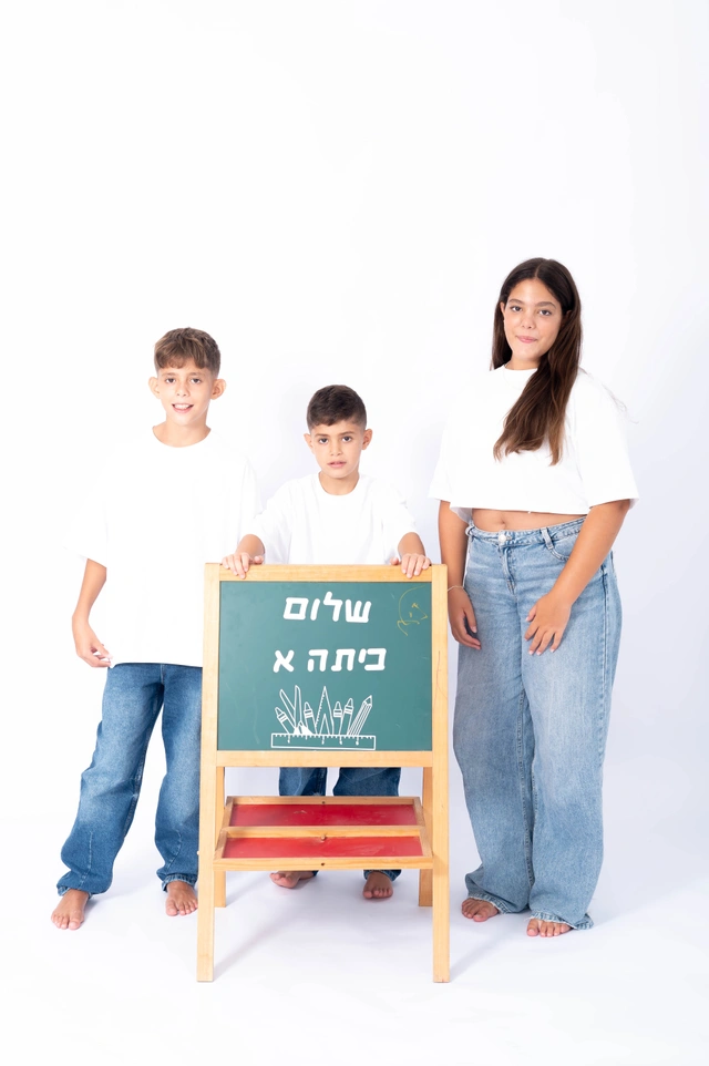 שלושה ילדים, שני בנים ובת, עומדים בסטודיו עם לוח גיר שעליו כתוב 'שלום כיתה א'' בעברית.