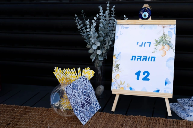שלט עץ קטן עם הכיתוב "רוני מרין 12" לצד כוס קש ופרחים