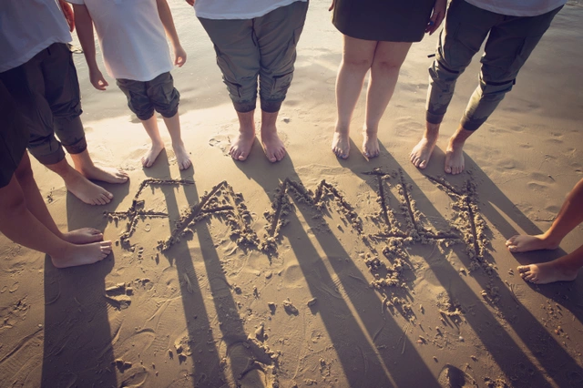 קבוצת אנשים עומדת יחפה על החוף מסביב למילה "FAMILY" הכתובה בחול.