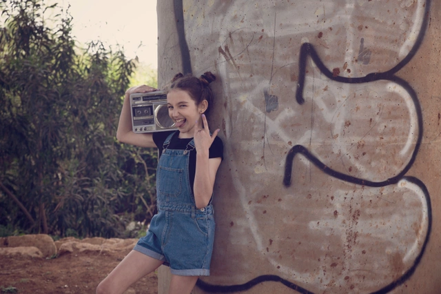 ילדה מצטלמת עם רשמקול, נשענת על עמוד גרפיטי.