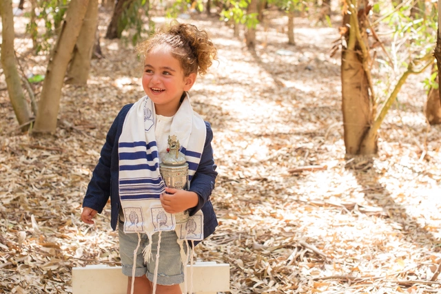 ילד מחייך בטלית מחזיק מעיל תורה קטן בחוץ.