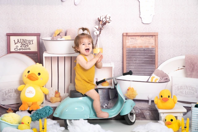 פעוטה מחייכת יושבת על קורקינט צעצוע בסטודיו בעיצוב חדר כביסה.