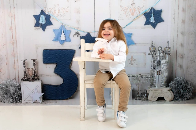 ילד בחולצה לבנה וכיפה יושב על כיסא וצוחק, על רקע עיטורי פורים.