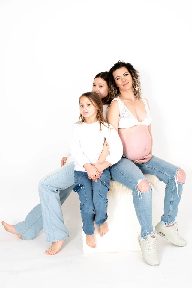 אישה בהריון עם שתי בנותיה יושבות יחד ומביטות למצלמה בסטודיו.