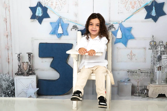 ילד מחייך יושב על כיסא לבן, מוקף בקישוטי חנוכה.