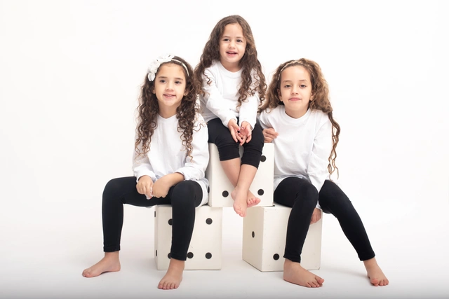 שלוש ילדות עם שיער מתולתל יושבות על קוביות לבנות בסטודיו.
