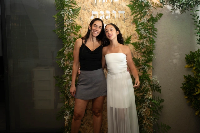 שתי נשים מחייכות ופוזות יחד מול רקע מעוטר בצמחייה.