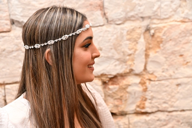 אישה מחייכת בפרופיל, עונדת סרט ראש מקושט, על רקע קיר אבן.